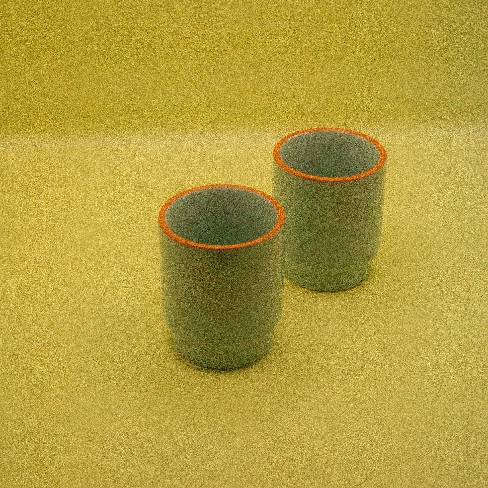 Two green ceramic cups with orange rims on a yellow background